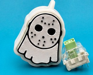 White 3D-printed ghost clicker keychain with a masked face, next to the mechanical switch that gives it its click